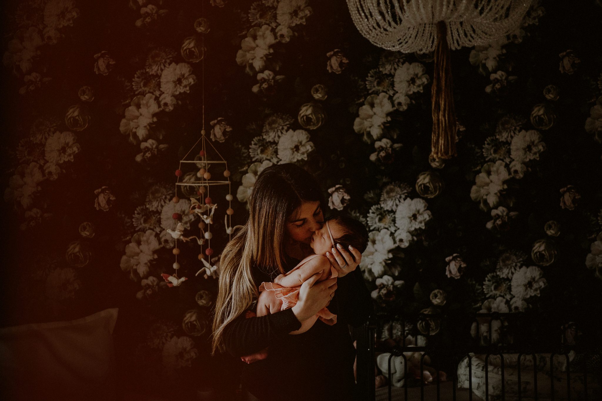 mother kissing baby in her arms in front of dark flowered wallpaper of bedroom