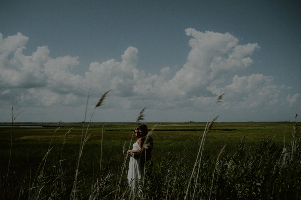 bride and groom wedding photo embraced among marshlands and clear blue sky with puffy white clouds - Bonnet Island Estate Wedding