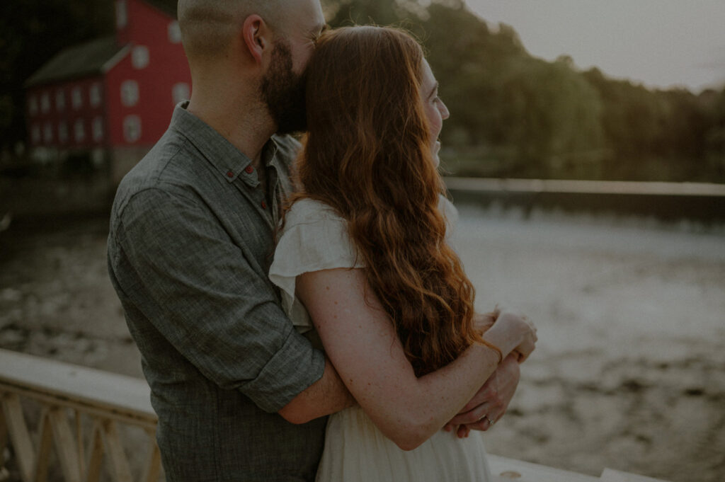 couple hugging in front of red mill museum in clinton new jersey overlooking river