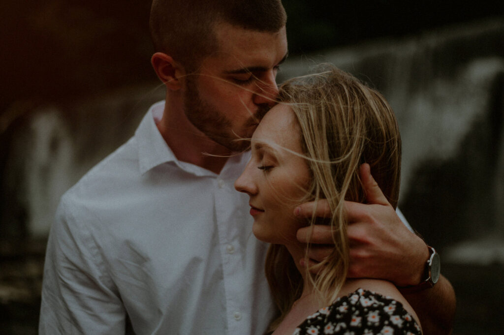 engagement photo of man kissing woman on the forehead in front of waterfall and grabbing her head 