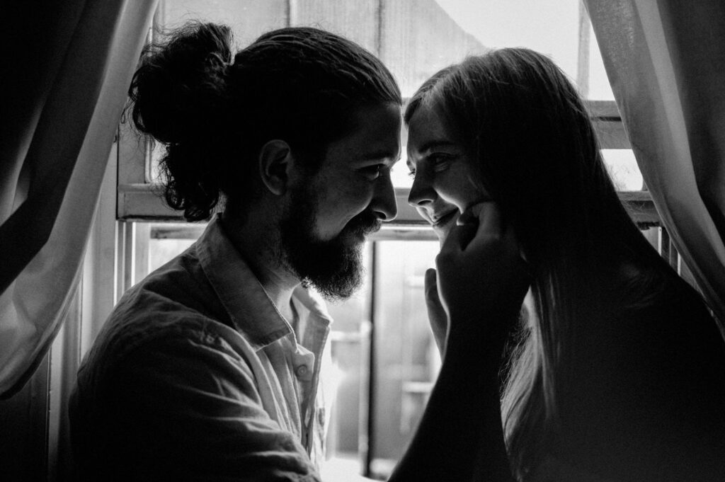 black and white photo of couple looking at each other while sitting on windowsill with hands on each other's faces
