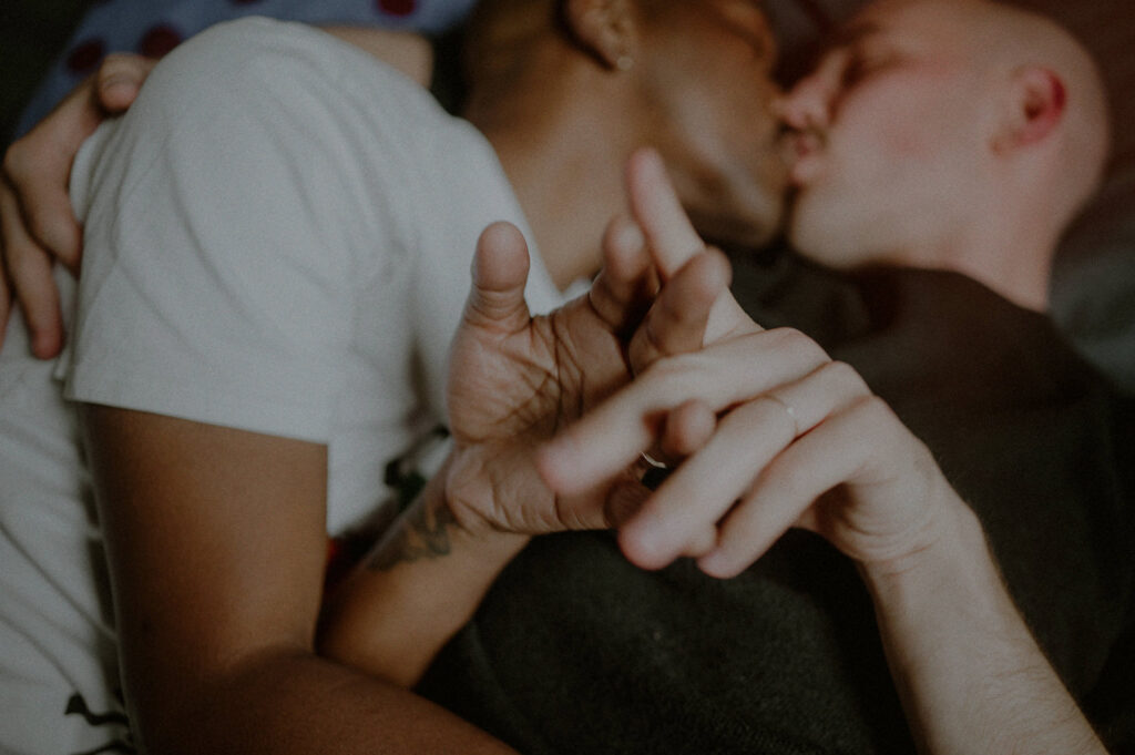 same sex couple laying in bed and kissing while playing with hands in foreground