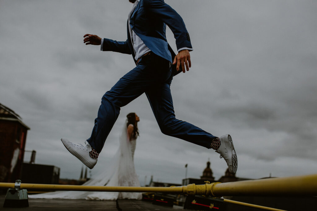 creative photo of groom jumping in air and bride in background through legs