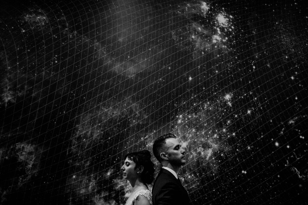 bride and groom posed back to back in black and white photo in front of a picture of the universe