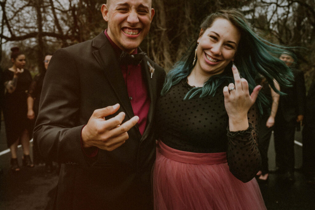 bride with pink dress and green hair and groom with red shirt and black suit laughing and posing with middle finger at camera