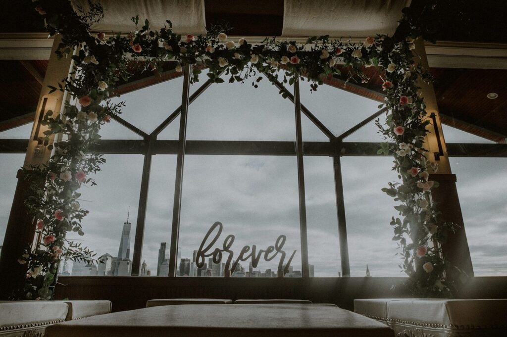 floral decorated mandap at liberty house indoor ceremony room overlooking nyc skyline with forever sign on window