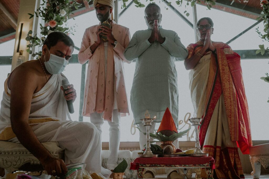 priest sits and prays as groom and family stand and pray in background