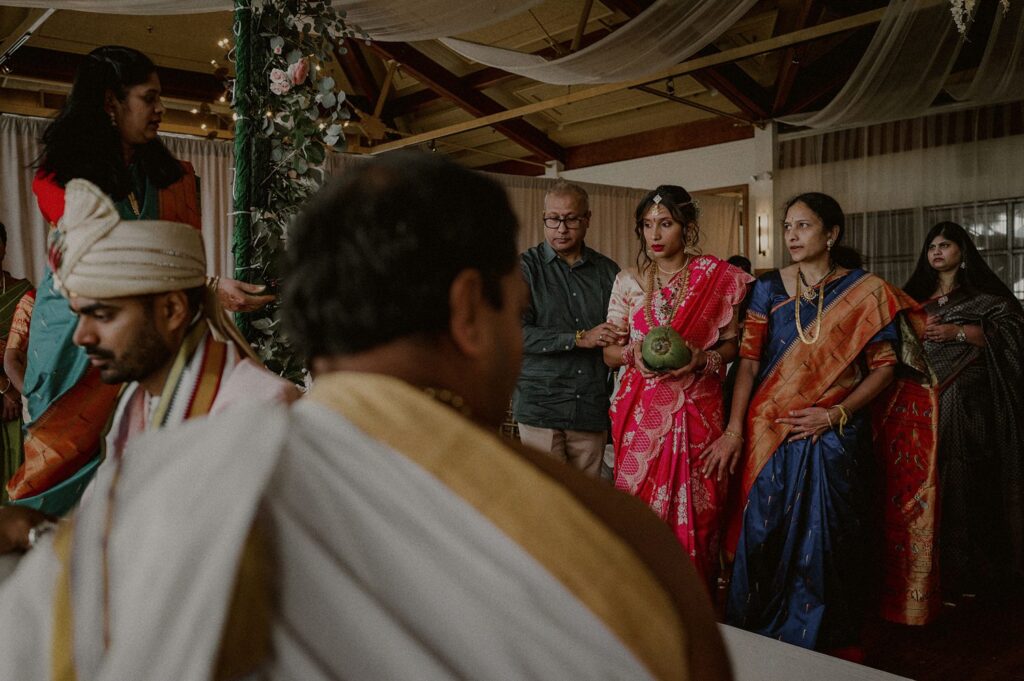 bride enters ceremony space mandap holding coconut with groom turned around