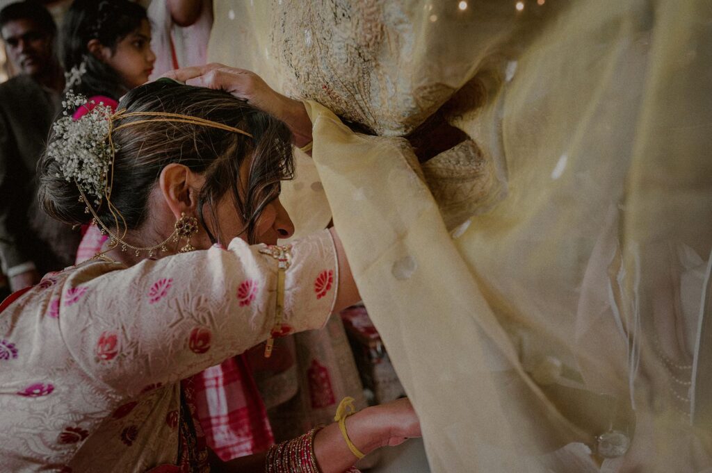 groom's hand on top of bride's head peeking out from yellow sheet as part of indian wedding tradition