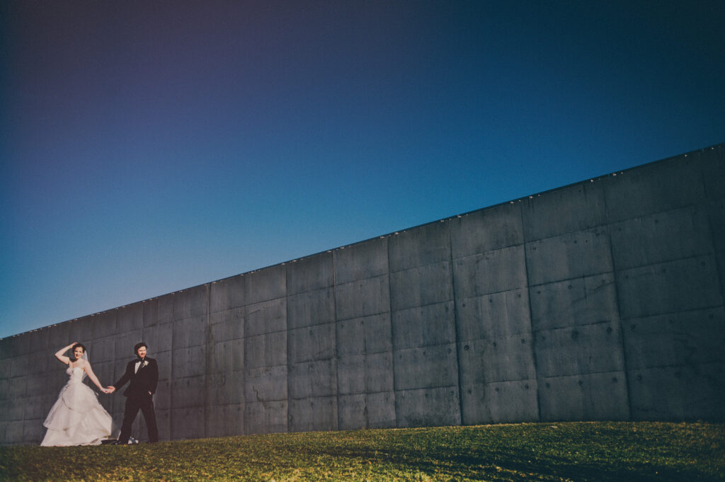 bride and groom walking off in casual portrait in front of wall at liberty state park