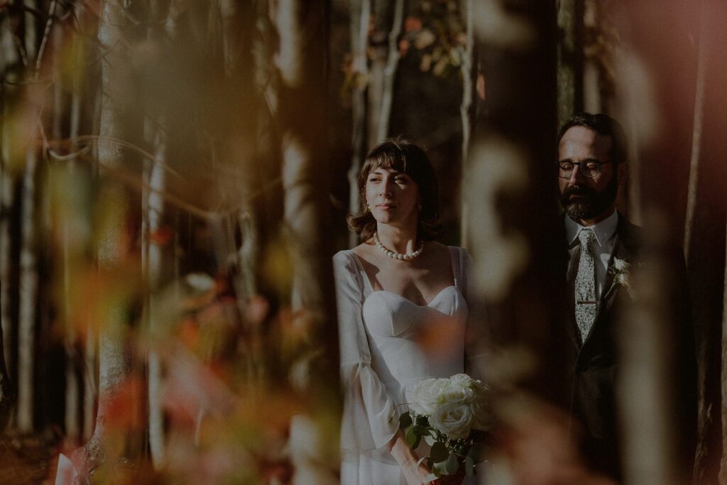moody portrait in dappled light of bride and groom posed between birch trees