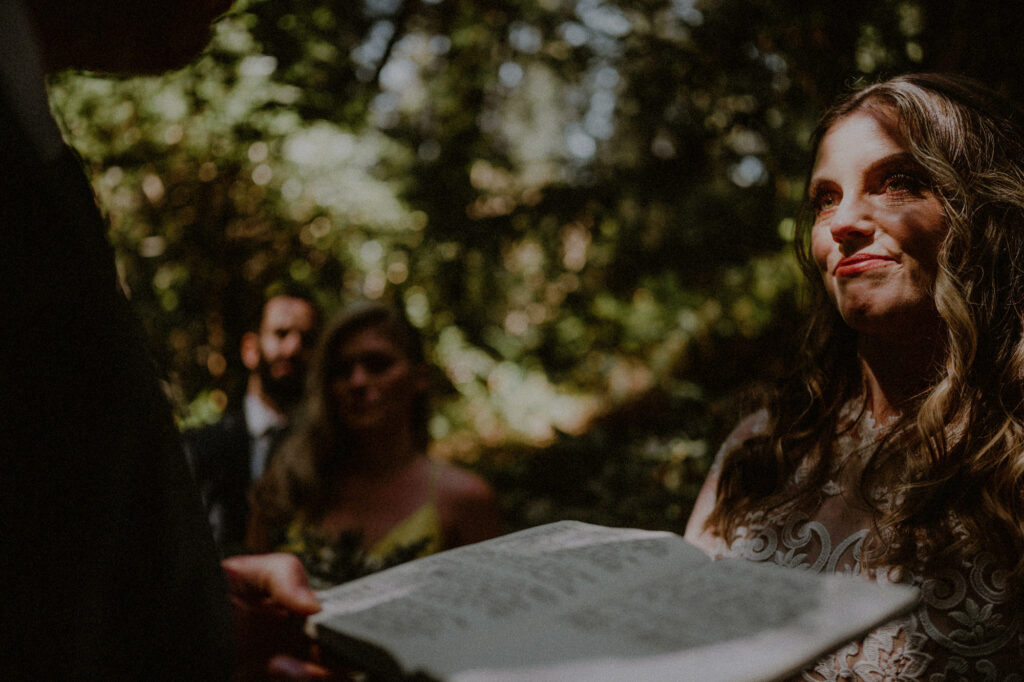 woman looking emotionally and framed with speckled sunlight as man reads his vows in the middle of the redwoods forest