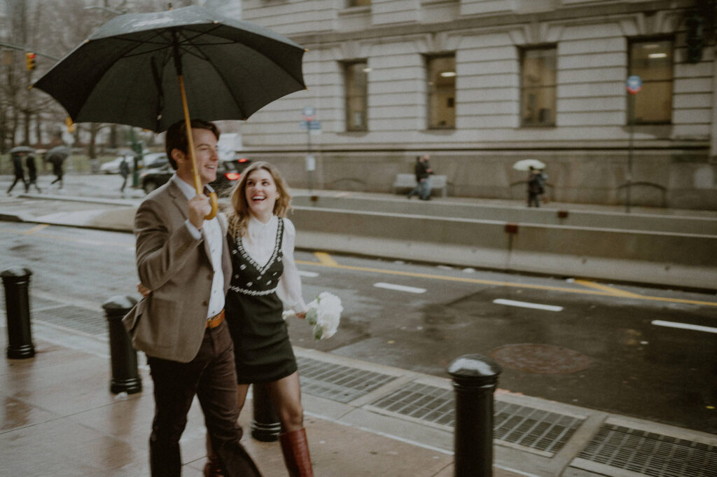 couple hastily walking in the nyc street with umbrella and laughing with each other to get to their elopement on time