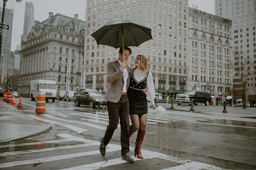 couple walking across downtown nyc street with umbrella in the rain after their city hall elopement